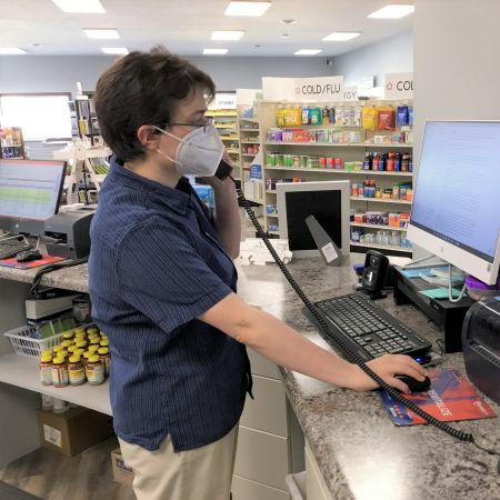 [Image description: A picture taken from the right side of Jay Holloway in profile standing at a pharmacy counter talking with a patient on the phone. He has short brown hair, and is wearing a dark blue shirt, khaki pants, glasses, and a white KN95 mask. He is looking at a black and white computer displaying the USCF Transgender Guidelines webpage. His phone is in his left hand, and the black cord stretches across to the right side of the counter.]