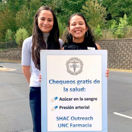 Student pharmacists Aliyah Cruz (right) and Belyin Gutierrez, co-directors of the Student Health Coalition Free Clinic at the UNC School of Pharmacy, hold a Spanish-language sign to announce free health checks, which were held outdoors during the COVID-19 pandemic.